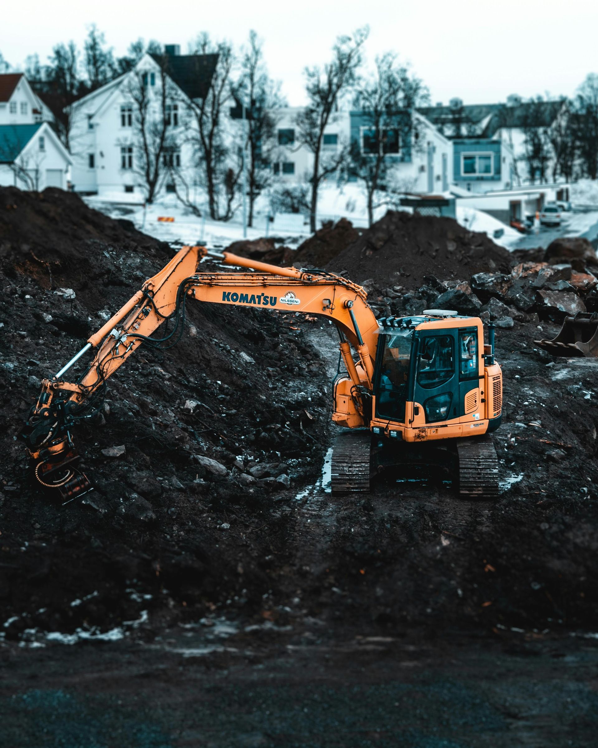 Yellow excavator on a mound of dark dirt, residential buildings in the background.