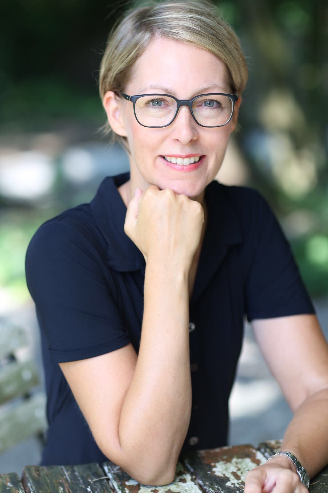 Woman in glasses smiling, resting chin on hand, wearing a black shirt, outdoors.