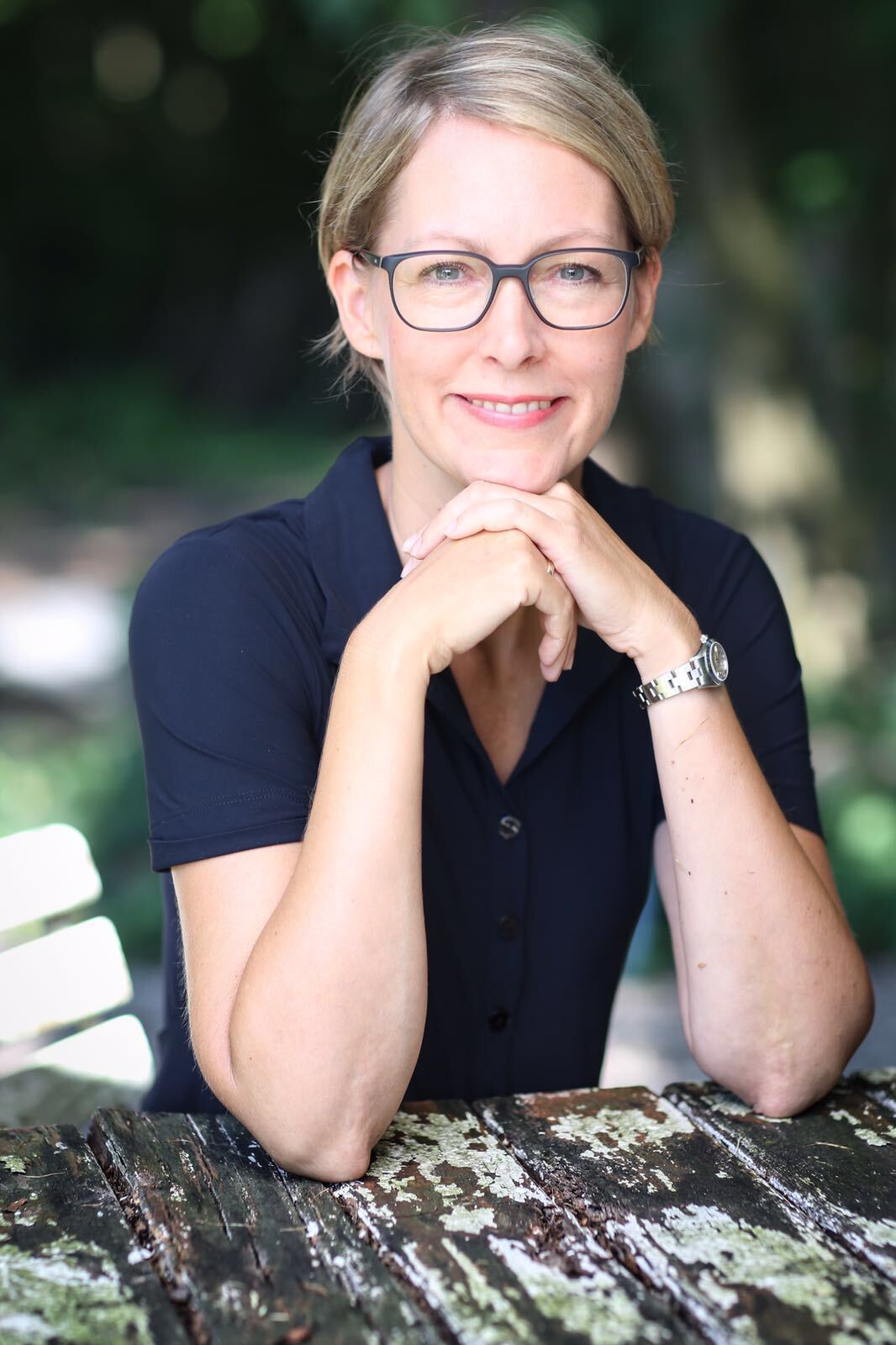 Woman in glasses resting chin on hands, outdoors at a weathered table, smiling.