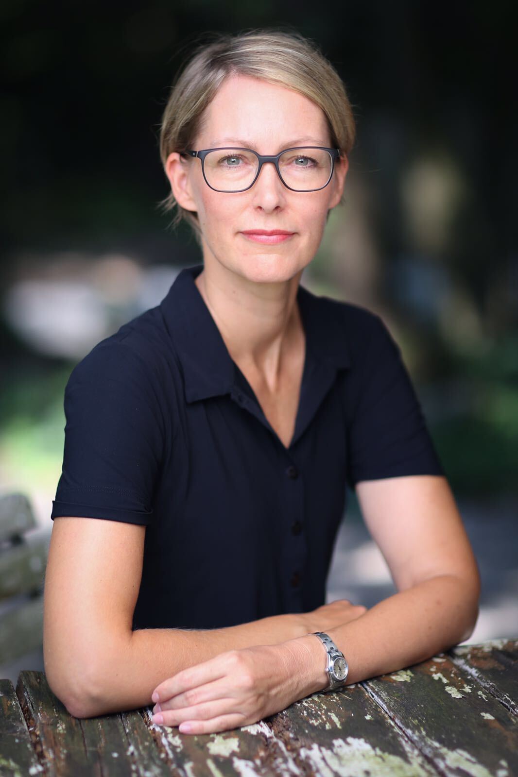 Woman with glasses, short blonde hair, wearing a dark blue shirt, arms crossed on a wooden table outdoors.