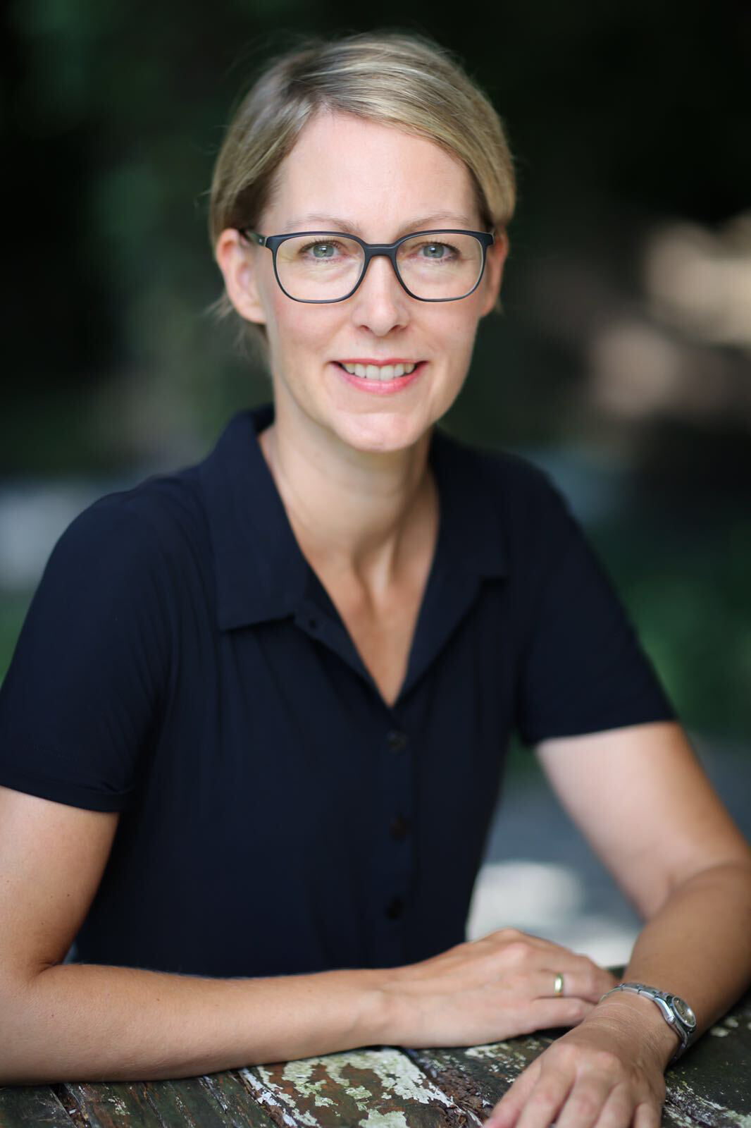 Woman with glasses smiles at the camera, wearing a dark blue shirt, sitting outside.