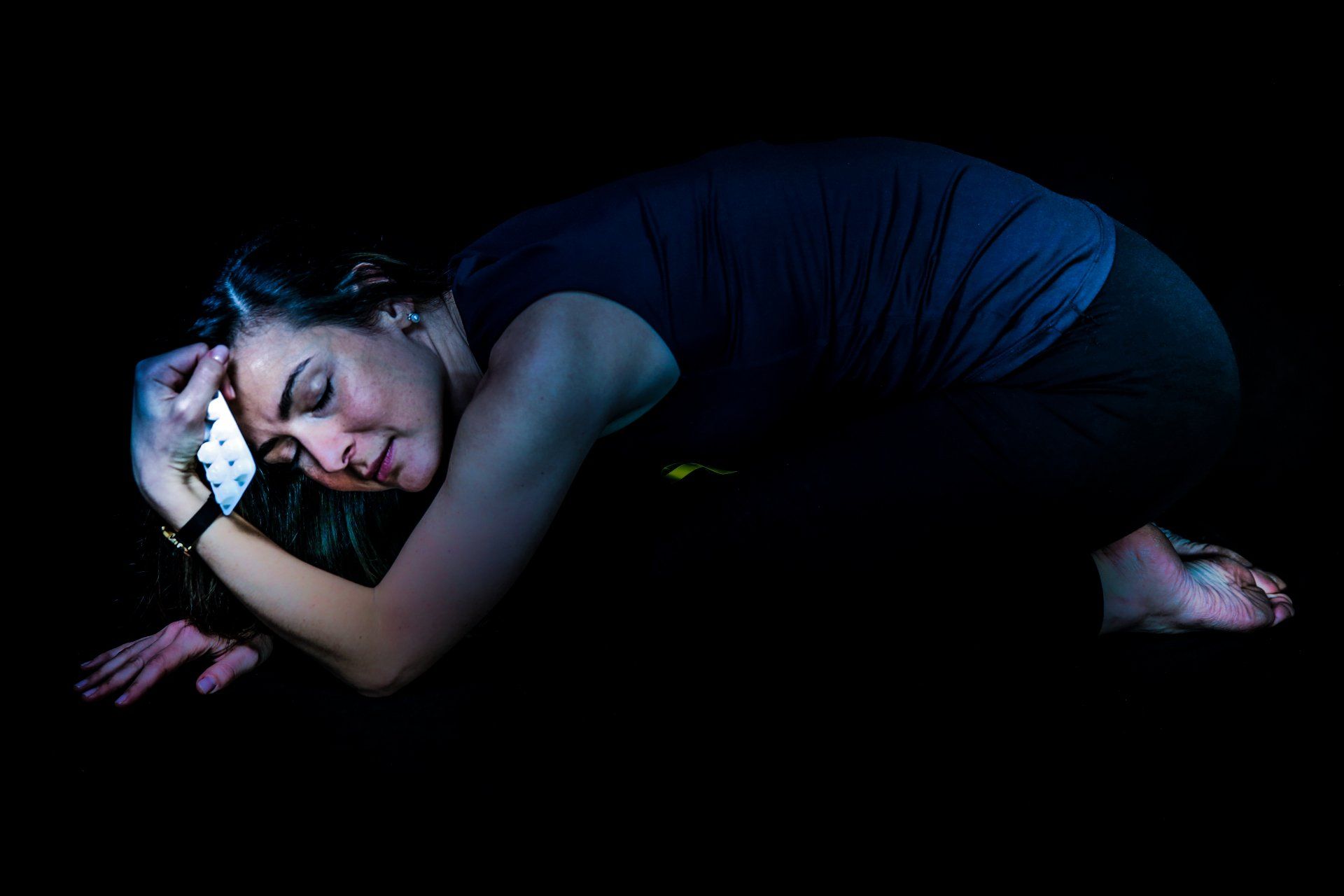 Woman hunched over, face in hand, in the dark; illuminated by blue and green light.