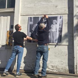 Two men hang a large black and white photo on a weathered, white garage door in sunlight.