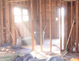 Sunlight streams into a gutted room, revealing wooden studs, debris, and tools like shovels and buckets.
