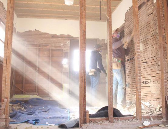 Men removing plaster from walls during a home renovation, with sunlight streaming through the room.