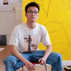Asian man with glasses sits on a red stool, wearing a t-shirt and jeans, yellow wall behind him.