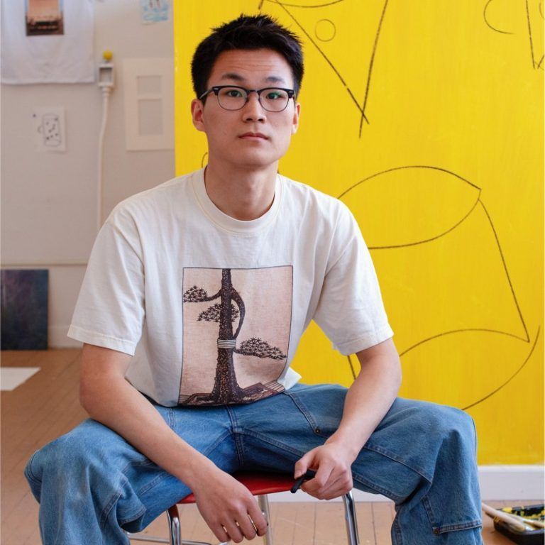 A young man wearing glasses is sitting on a chair in front of a yellow wall.