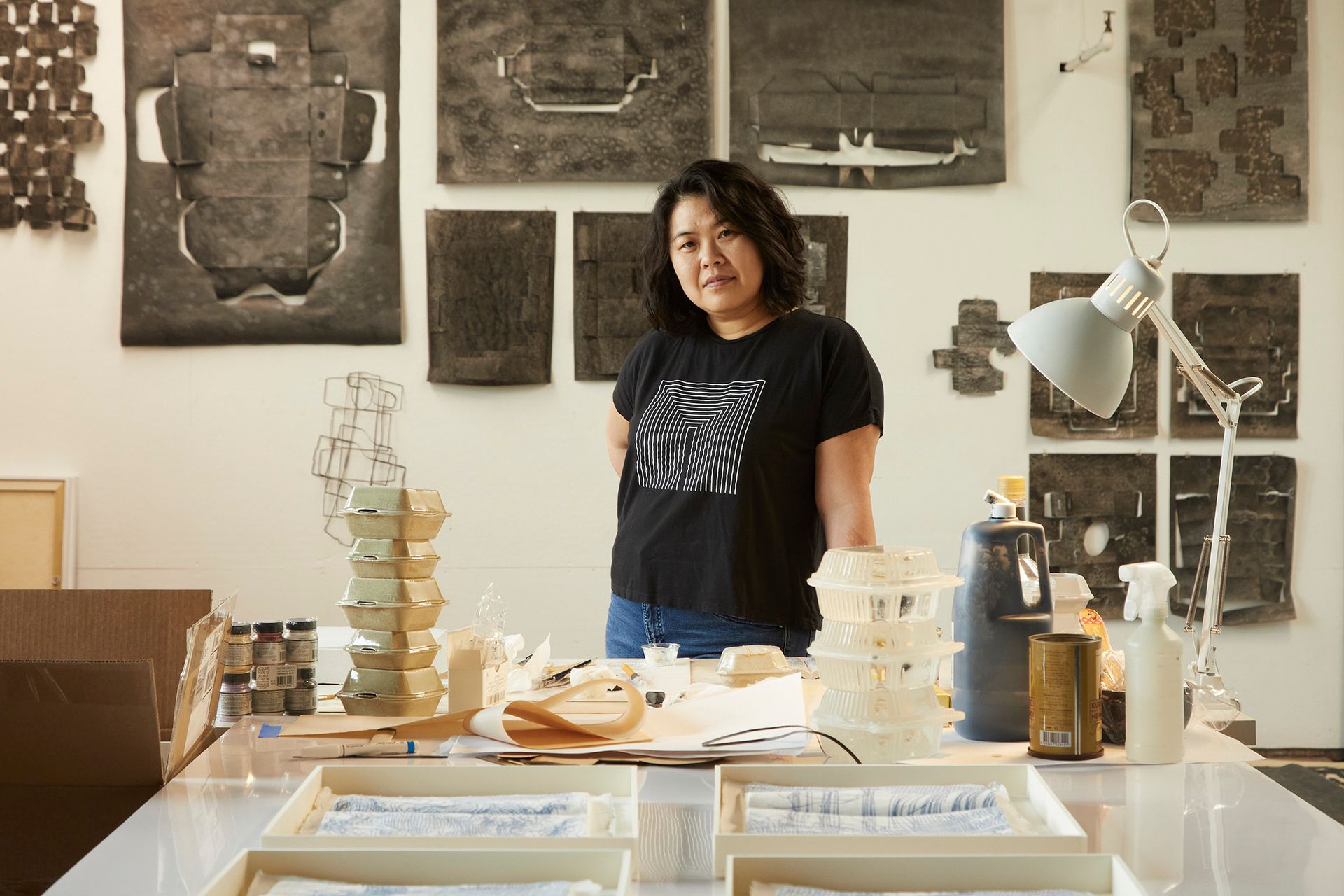 Artist standing at a table in a studio, surrounded by artwork.
