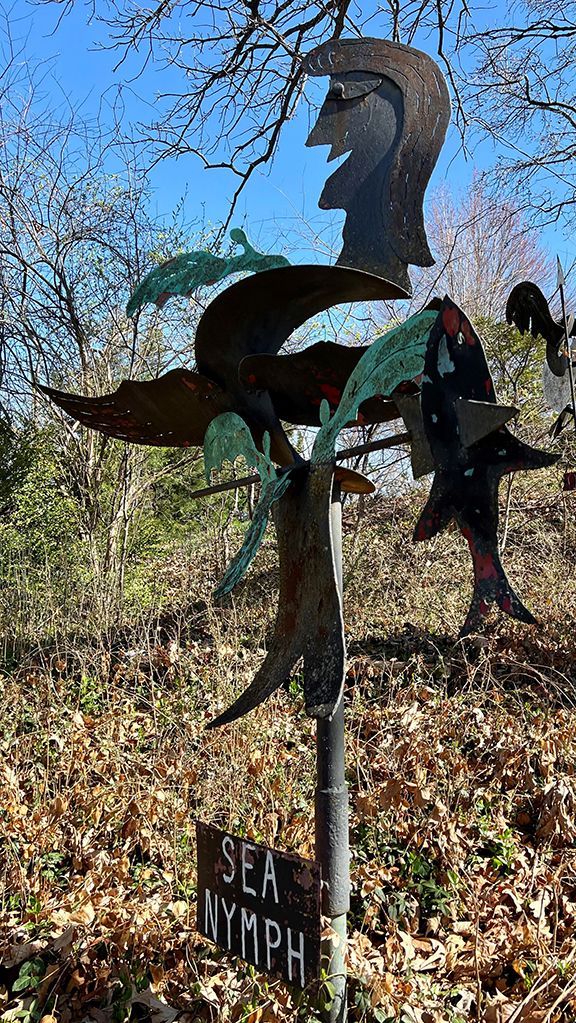 Metal sculpture of a sea nymph, standing in a wooded area with a blue sky background.
