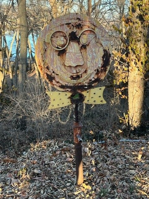 Rusty metal sculpture of a face with glasses and bow tie, in a wooded area.