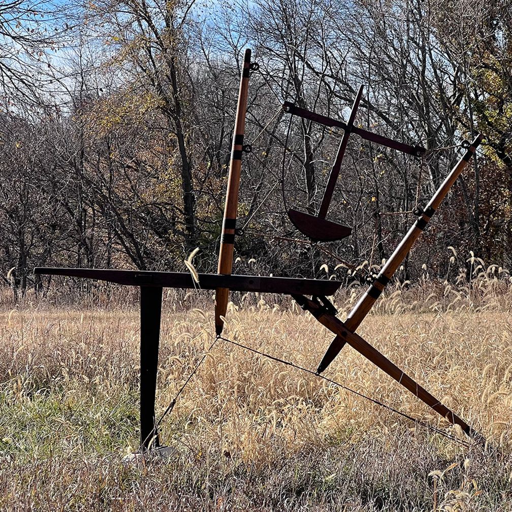 Abstract outdoor sculpture in a field. Brown and black metal structures against a backdrop of trees and dry grass.
