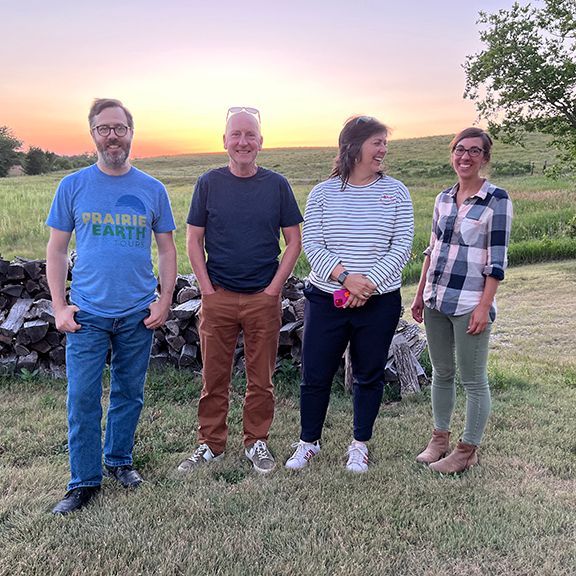 Four people stand in a field at sunset. A man in blue smiles. Woman in stripes laughs. Others look on.