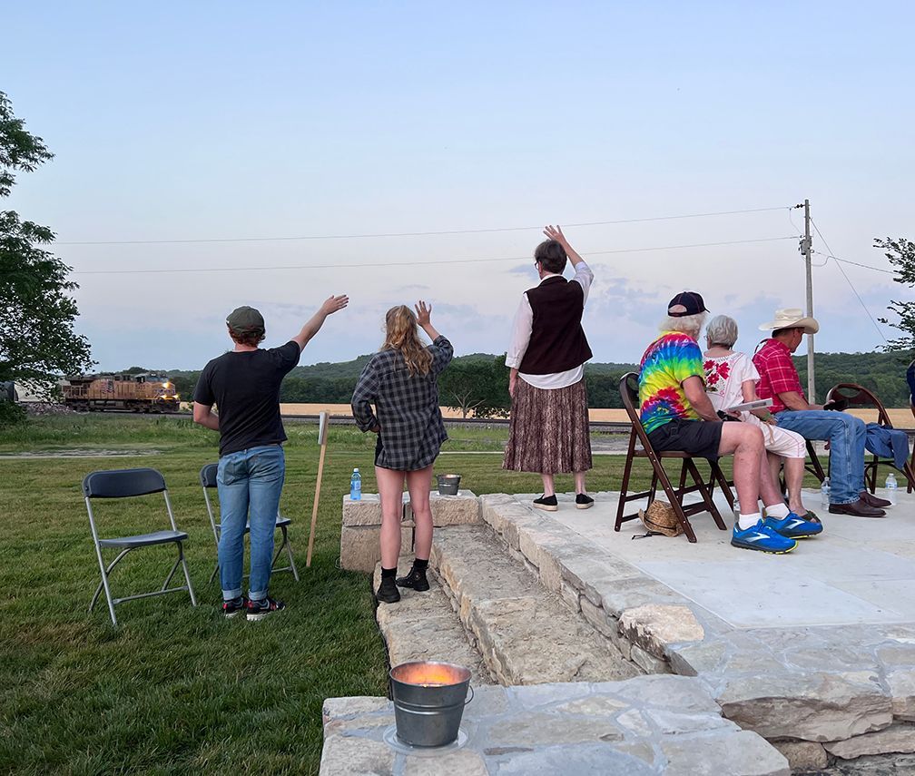 People wave at a passing train from an outdoor gathering at dusk.
