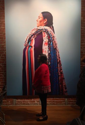 Woman in red sweater stands before a large portrait of a Native American woman in traditional clothing.