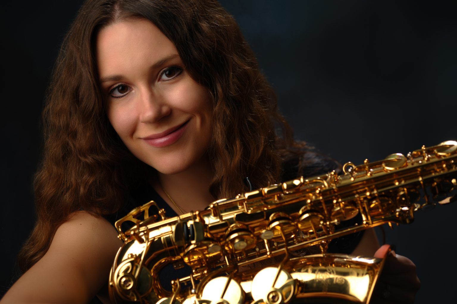 Woman with long brown hair holding a golden saxophone, smiling against a dark background.