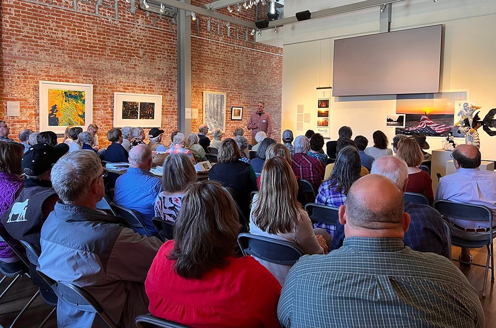 Audience seated in gallery listening to a speaker, brick wall, artwork displayed, screen overhead.