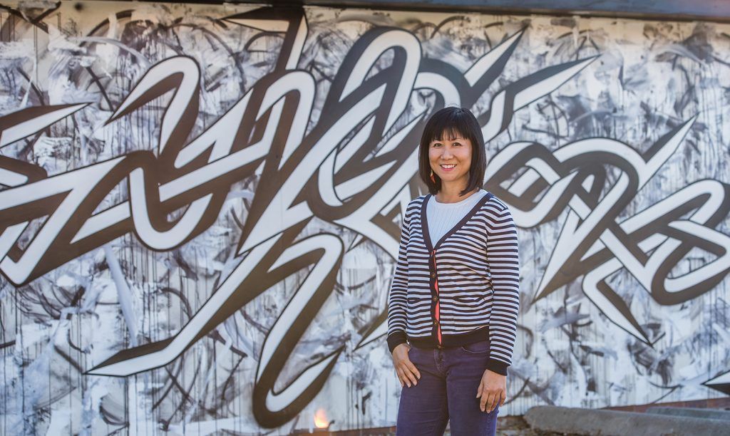Woman in striped jacket smiles in front of a black and white graffiti mural.