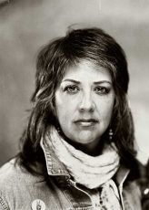 Woman with short wavy hair, wearing denim jacket and scarf, looking straight ahead.