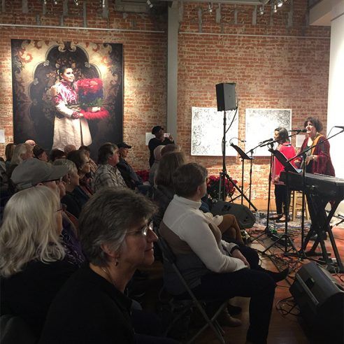 A live music performance at an art gallery. Two women sing on stage; audience seated, watching, and listening.