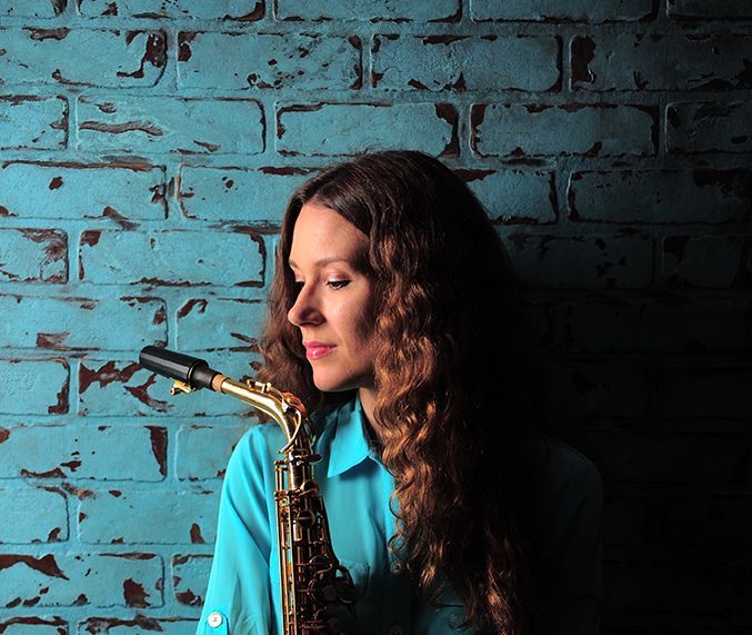 A woman in a blue shirt is playing a saxophone in front of a blue brick wall.