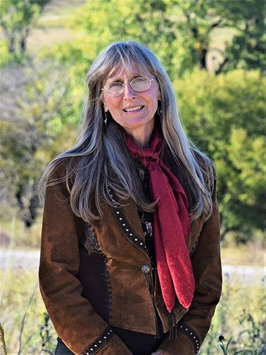 Woman with long gray hair smiles, wearing a brown suede jacket and red scarf, outdoors.