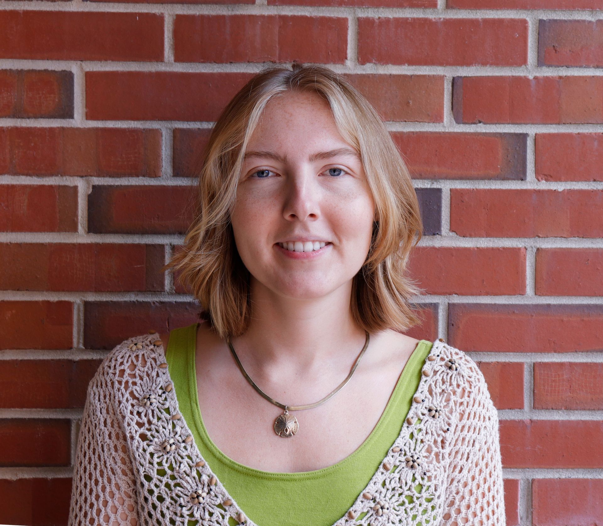 Woman with blonde hair smiling in front of a brick wall, wearing a green shirt and a crochet sweater.