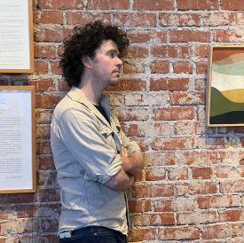 Man with curly hair, arms crossed, stands in front of a brick wall with framed art.