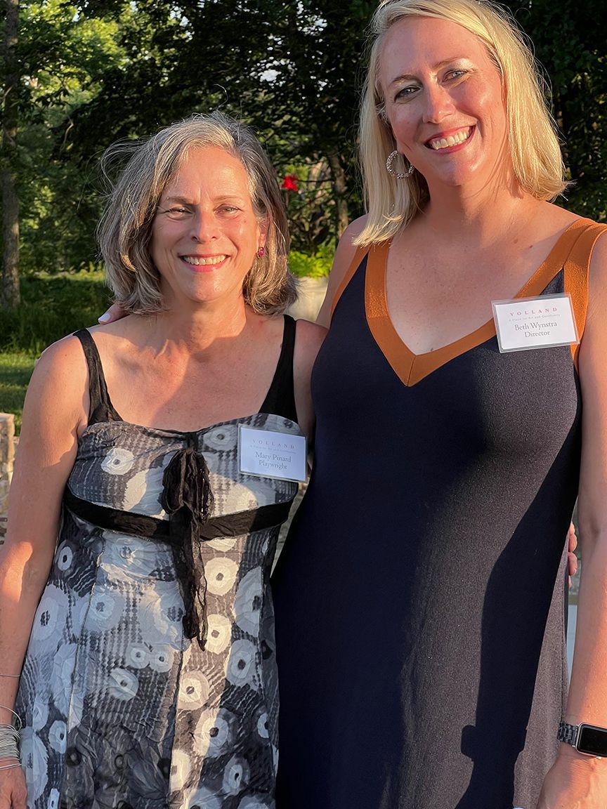 Two smiling women pose outdoors. One in a floral print dress, the other in a blue dress with tan trim.