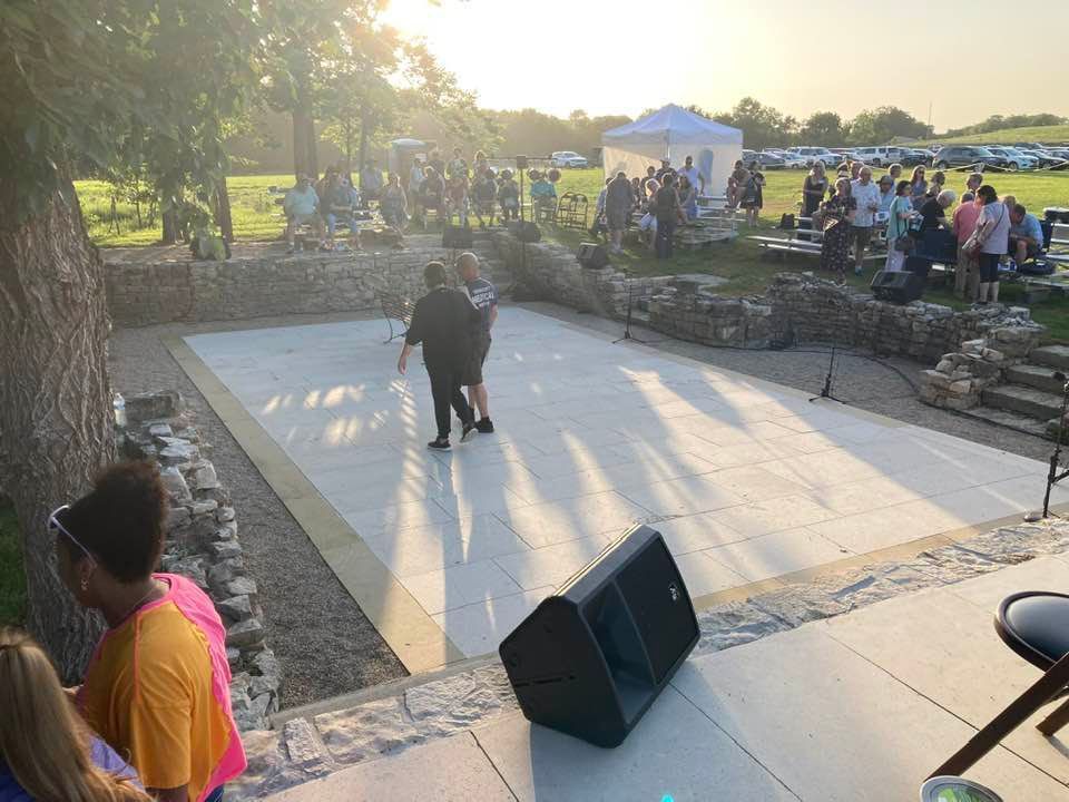 People dance on a stage at an outdoor event, with an audience and sunset in the background.