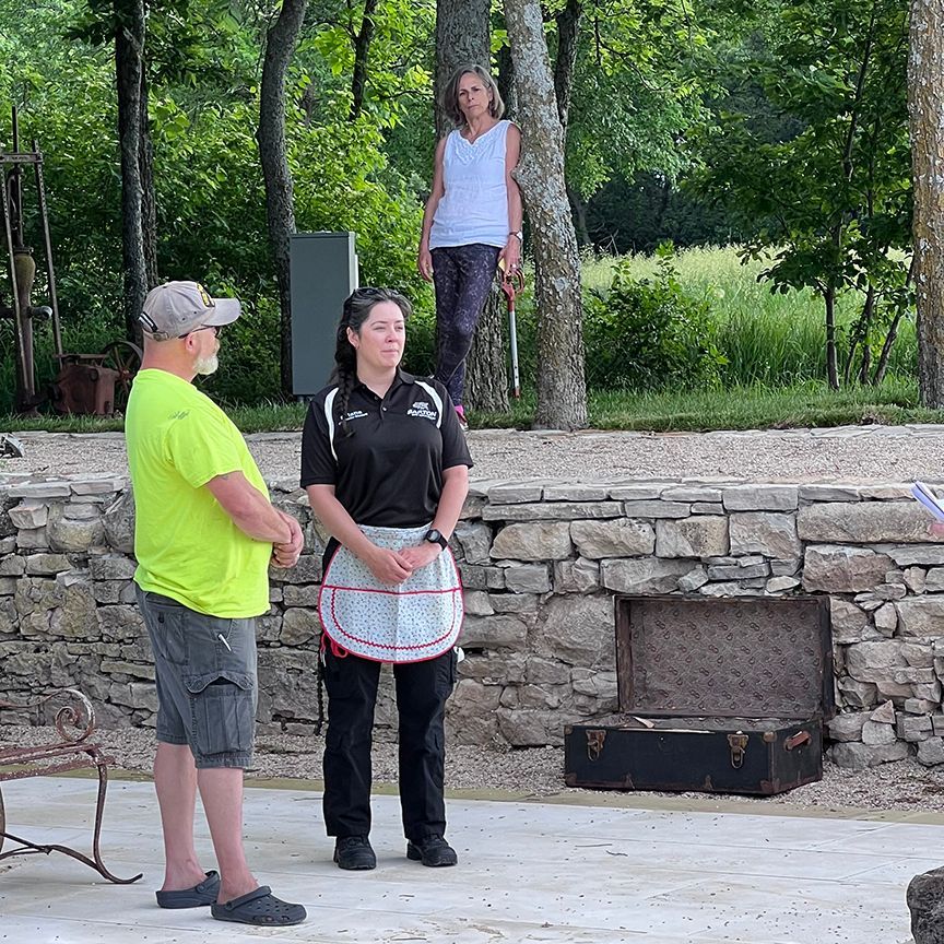 Three people outdoors: a man in yellow shirt and shorts, a woman in black uniform and apron, and another woman standing near a tree.
