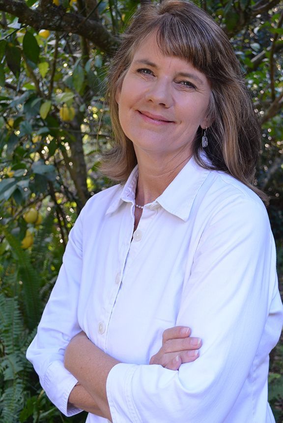 A woman in a white shirt is standing with her arms crossed in front of a tree.