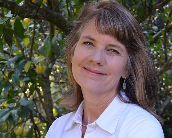 A woman in a white shirt is standing with her arms crossed in front of a tree.