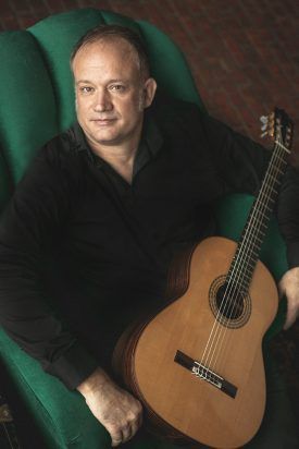Man in black shirt sits, holding acoustic guitar; emerald green chair, warm lighting.