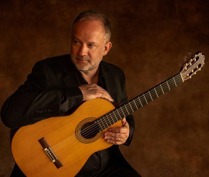 Man with guitar, looking thoughtfully away, in a dimly lit studio. He wears a black suit, brown background.