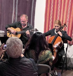 Guitarist and cellist perform; audience watches in a room with patterned curtains.