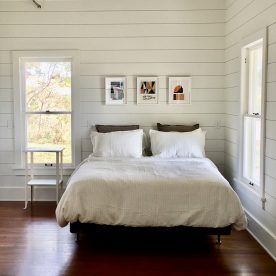 Bedroom with white shiplap walls, a bed, and three framed art prints. Sunlight streams in from two windows.