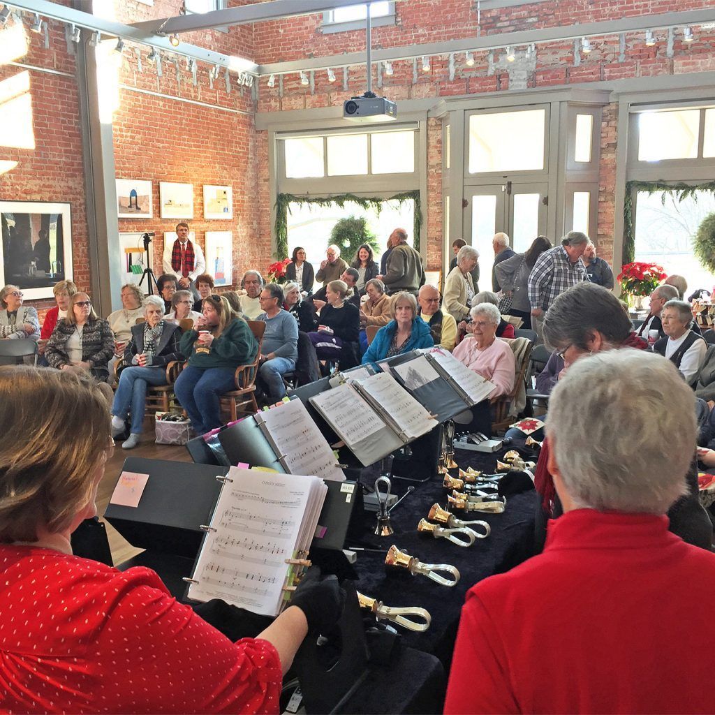 Handbell choir performing in a brick-walled room. Audience watches.