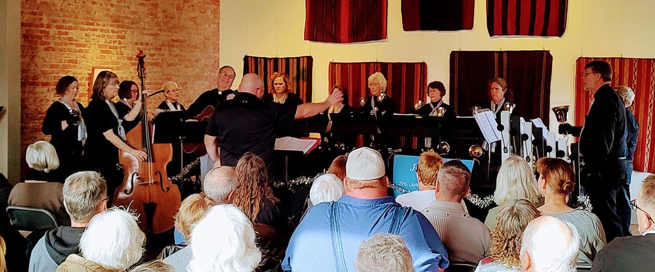 A musical performance with a band on a stage, audience seated. The setting is indoors, with a brick wall.