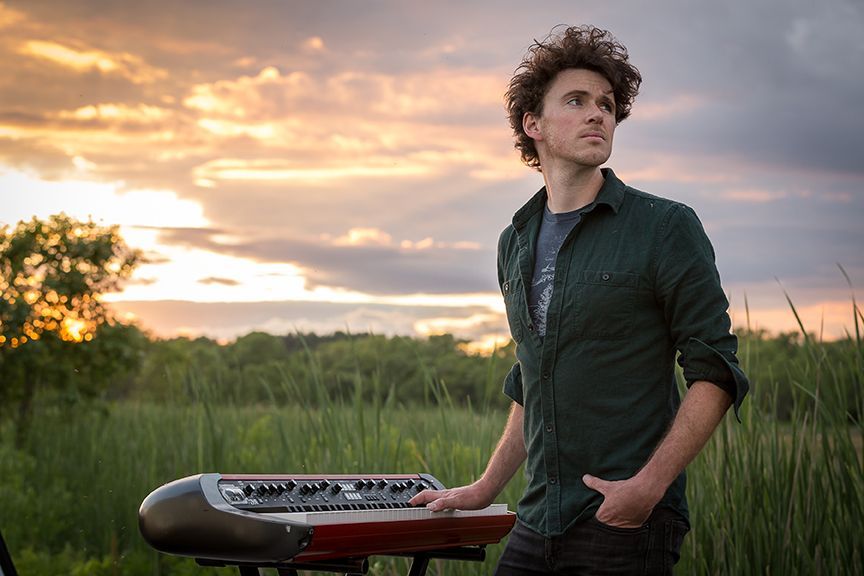 Man with curly hair standing near a keyboard, looking up at a sunset over a marsh.