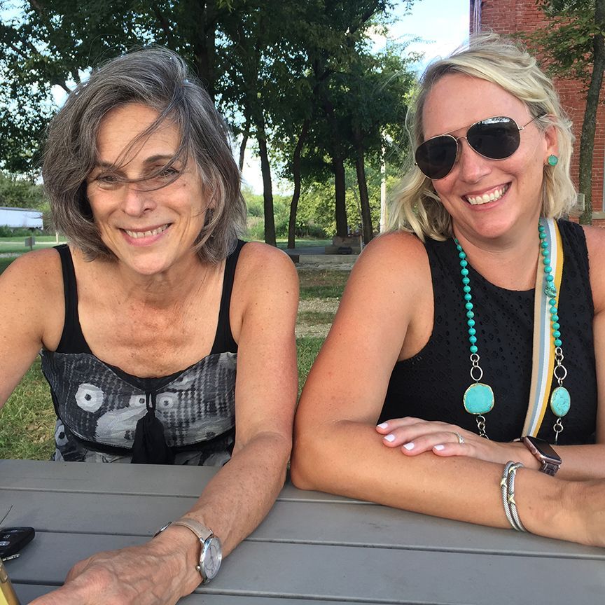 Two women smiling outdoors at a table. One has gray hair and a patterned top, the other blonde with sunglasses and turquoise jewelry.