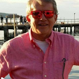 Man in red sunglasses and pink shirt smiles near a pier.