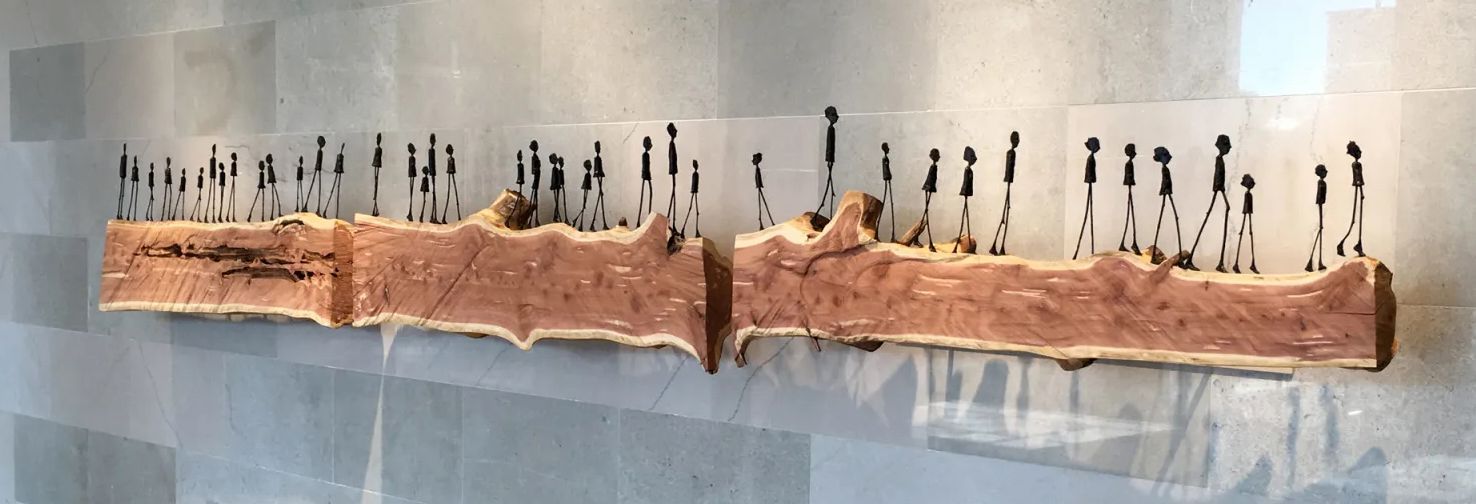 Art installation: silhouettes of people walking atop a wood sculpture against a wall.