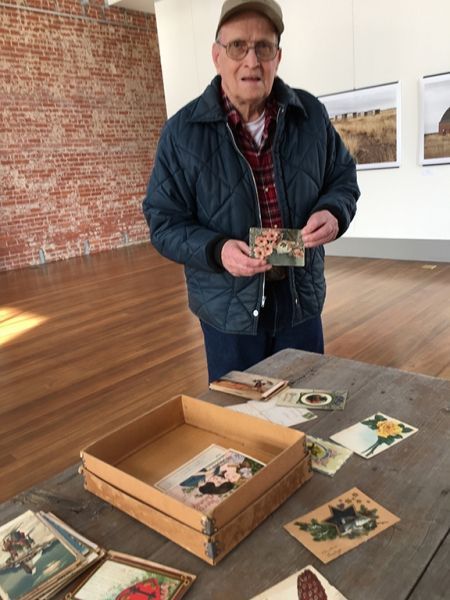 A man in a blue jacket is holding a postcard in front of a table full of postcards