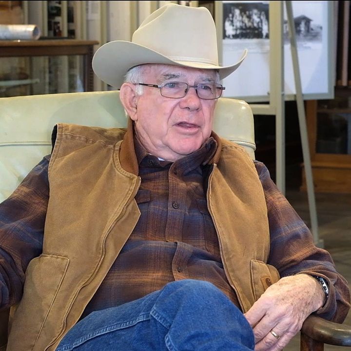 Older man in cowboy hat, vest, and glasses, seated indoors.