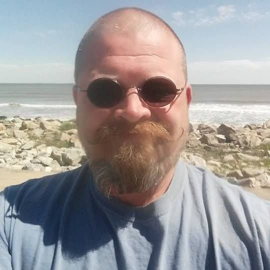 Man with beard and sunglasses at the beach, looking at the camera.