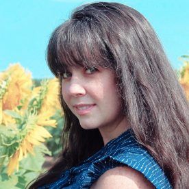 Woman smiling in front of sunflowers, wearing a blue shirt, against a blue sky.