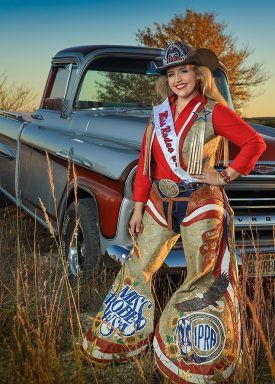 Woman in rodeo attire stands by vintage truck in field.