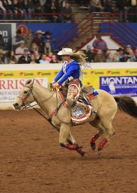 Cowgirl on tan horse in rodeo arena, wearing blue shirt, red chaps, competing.
