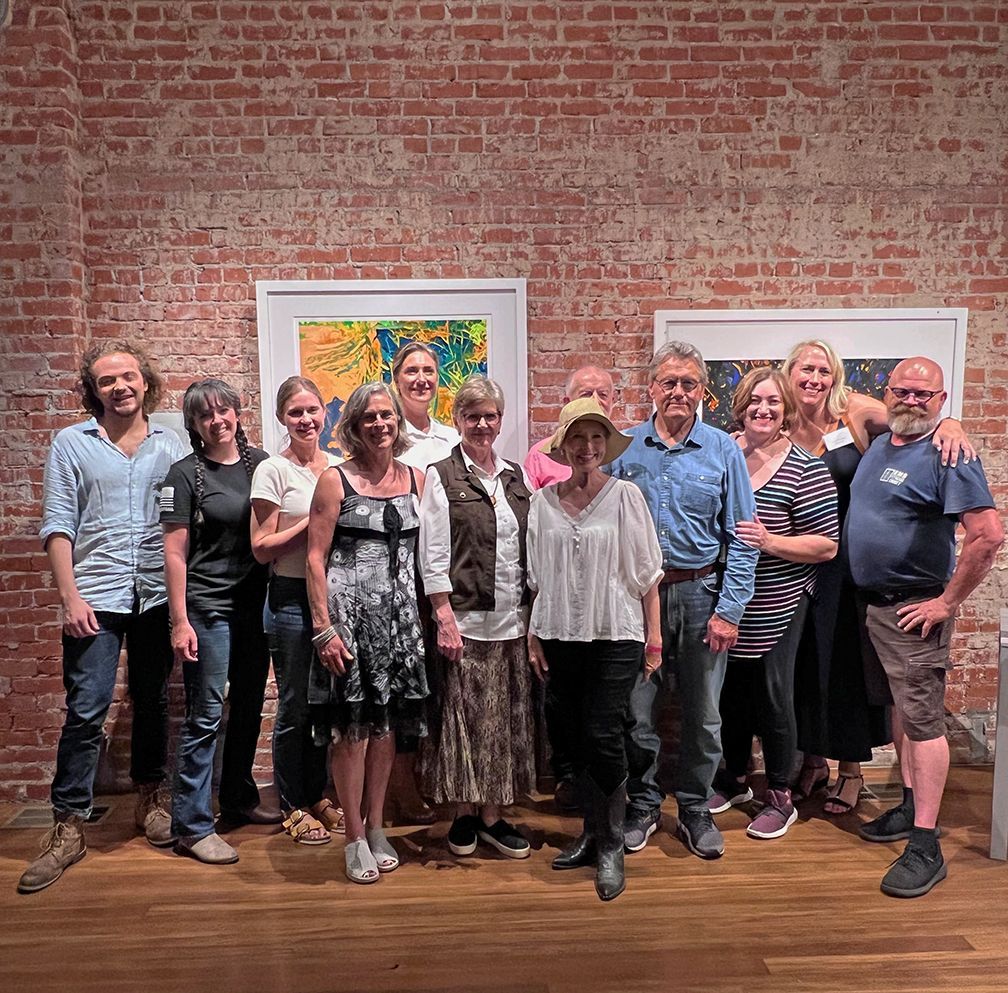 Group of people standing in front of paintings on a brick wall in a gallery setting.