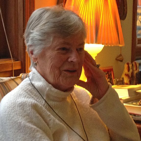 Elderly woman with short gray hair, wearing a white sweater, sits near a lamp.